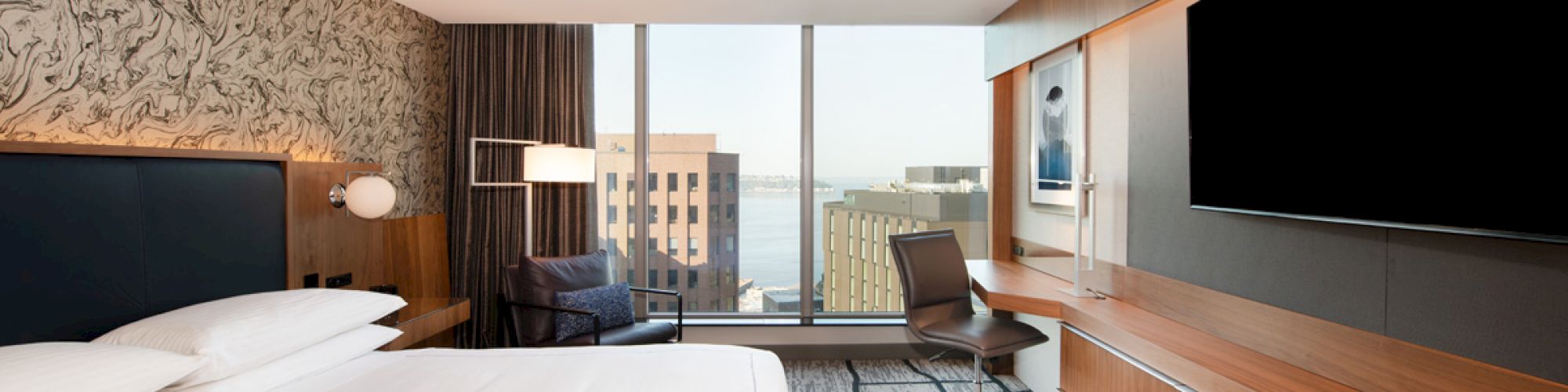 A modern hotel room with a large bed, desk, chair, big TV, and window overlooking city buildings.