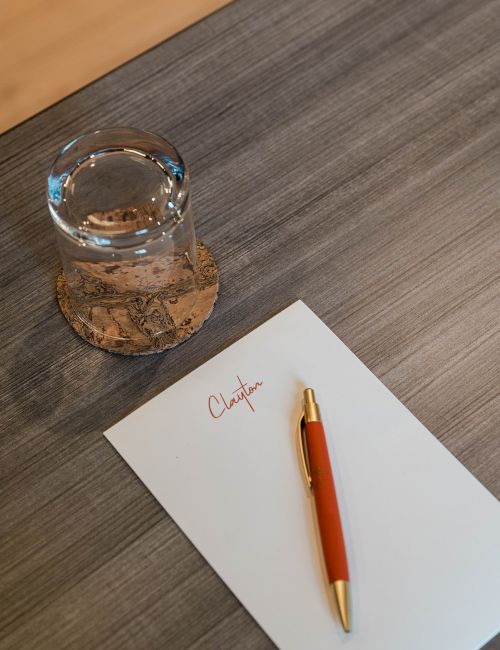 A glass paperweight sits beside a cork, with a red-gold pen and a blank white card on a wooden desk, ready for a note.
