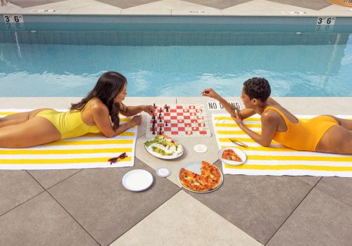 Two people in swimsuits lie on poolside towels facing each other, sharing pizza, salad, and drinks with a checkerboard napkin and sunlit pool backdrop.
