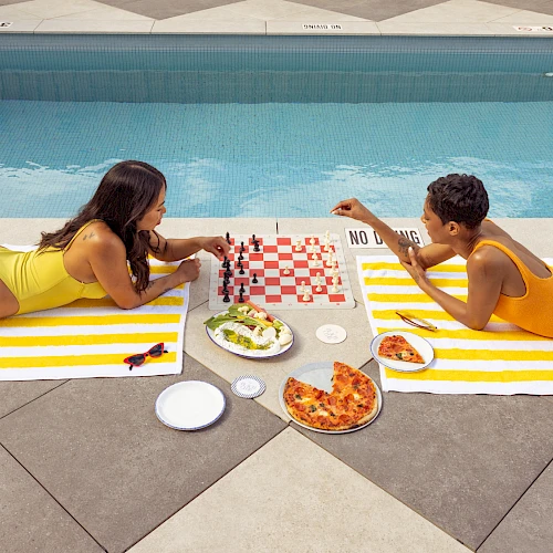 Two people in swimsuits lie on poolside towels facing each other, sharing pizza, salad, and drinks with a checkerboard napkin and sunlit pool backdrop.