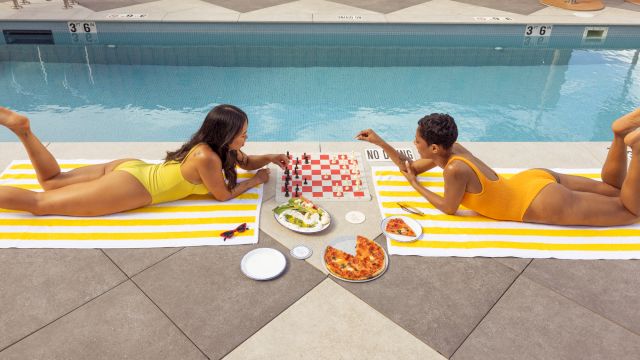 Two people in swimsuits lie on poolside towels facing each other, sharing pizza, salad, and drinks with a checkerboard napkin and sunlit pool backdrop.
