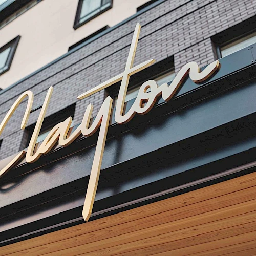 A stylish storefront with a gold cursive sign that reads &ldquo;Clayton&rdquo; against a brick-and-wood facade, under an overhang.